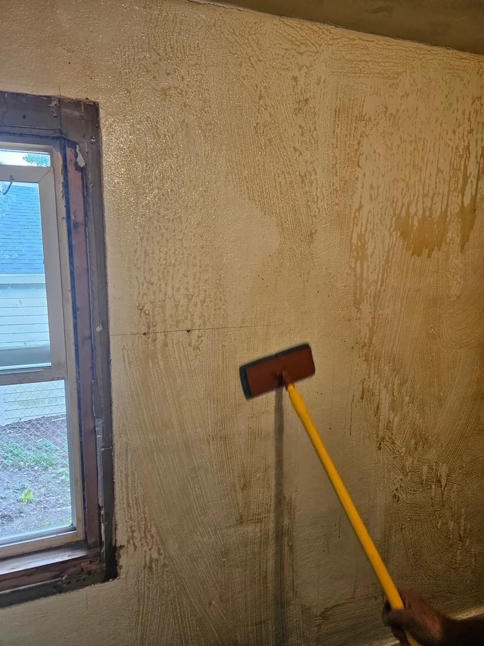 Cleaning pad on a yellow extension handle against a wall with brown residue and wet streaks beside a window